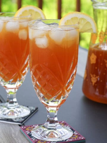 two crystal glasses of lemonade iced tea with lemon garnish and tea pitcher in background.